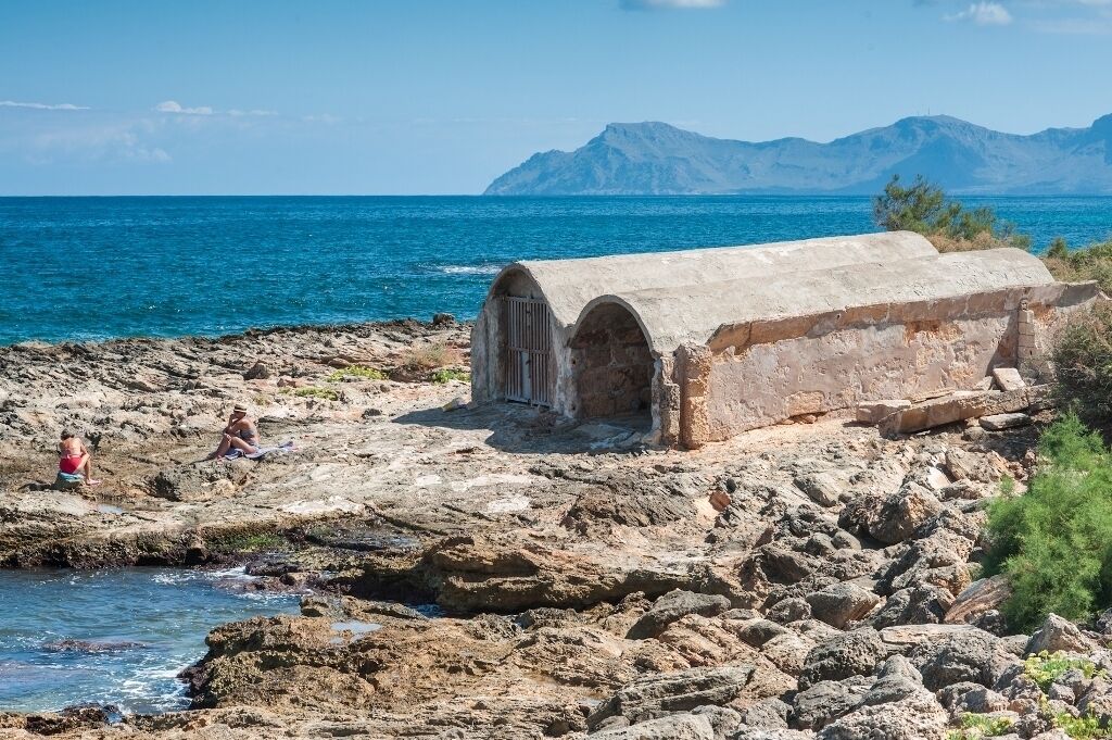 A stone structure with arched openings near a rocky shoreline and the sea.