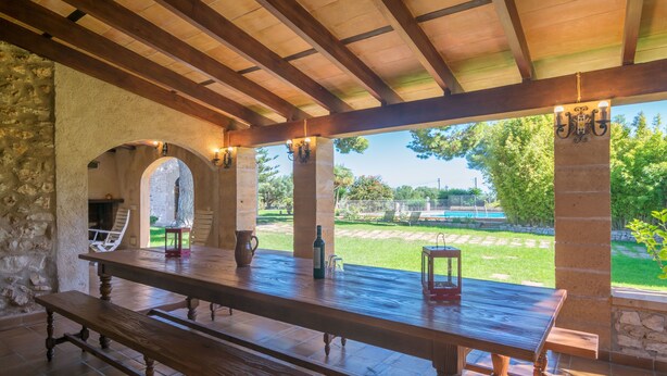 Eine Terrasse im Freien mit einem Holztisch und Bänken, Steinmauern und Blick auf Pool und Garten.