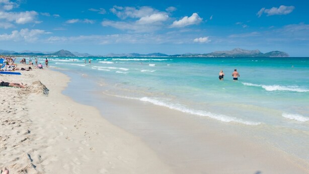 Ein Strand mit klarem türkisblauem Wasser, Menschen beim Schwimmen und Sonnenbaden, und Berge im Hintergrund.