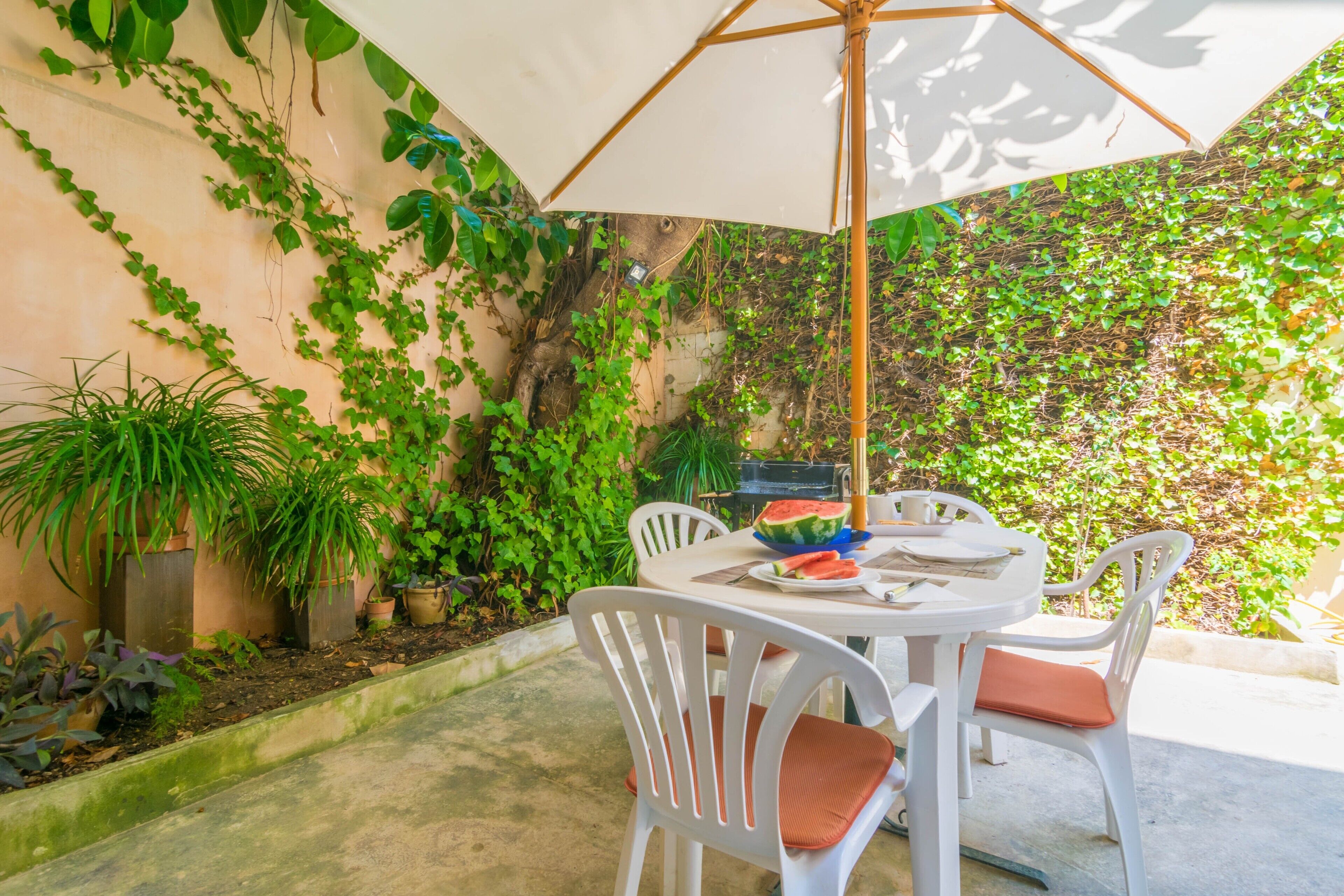 Eine Terrasse mit Tisch und Stühlen, umgeben von üppigem Grün und einem weißen Sonnenschirm.