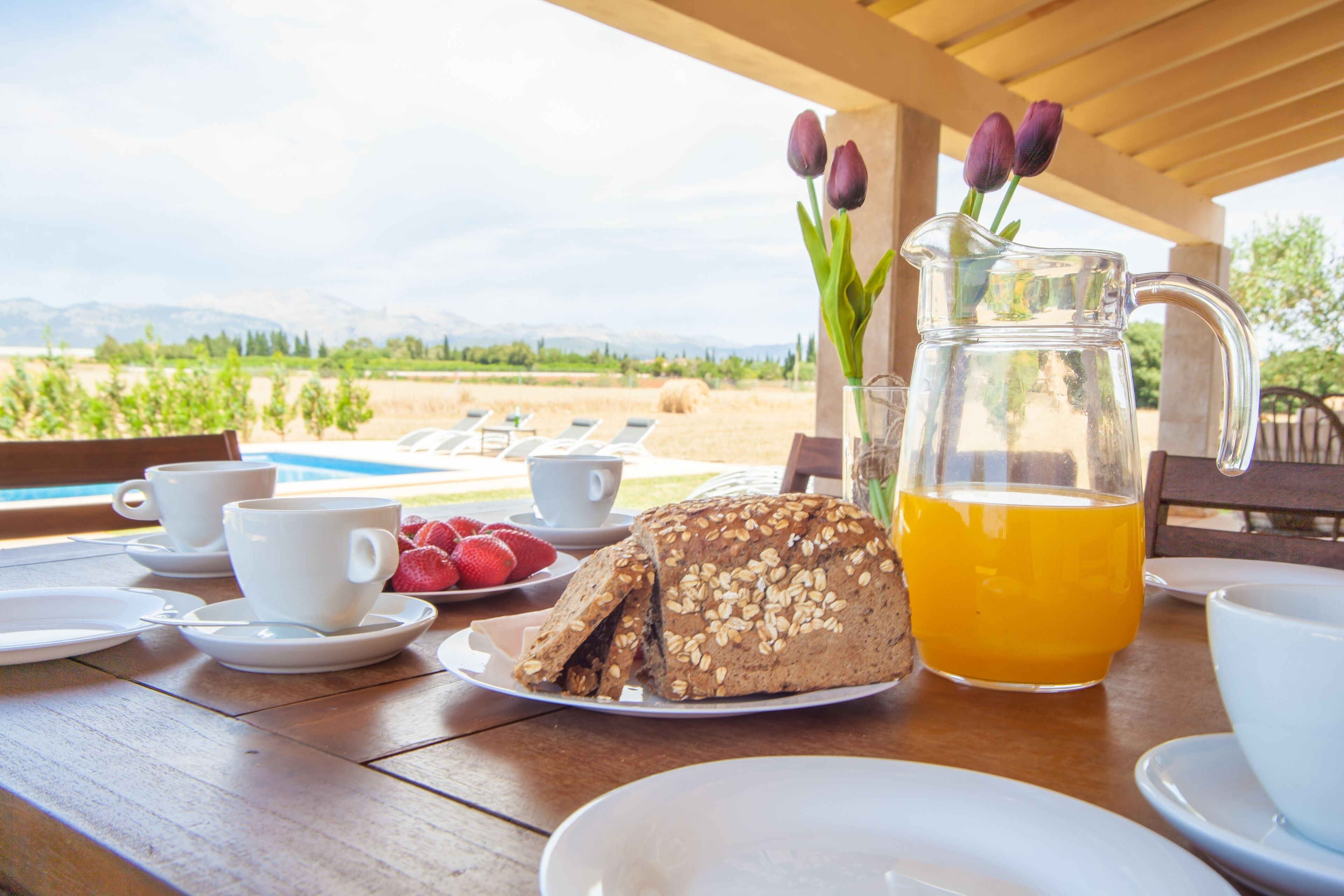 A breakfast table with a loaf of bread, a pitcher of orange juice, and a vase with tulips.