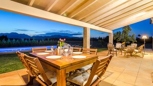 Eine Terrasse im Freien mit einem Holztisch, gedeckt für ein Essen, umgeben von Korbstühlen und Blick auf die Berge in der Abenddämmerung.