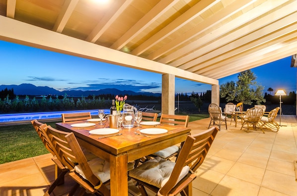 An outdoor patio with a wooden table set for dining, surrounded by wicker chairs, and a view of mountains at dusk.