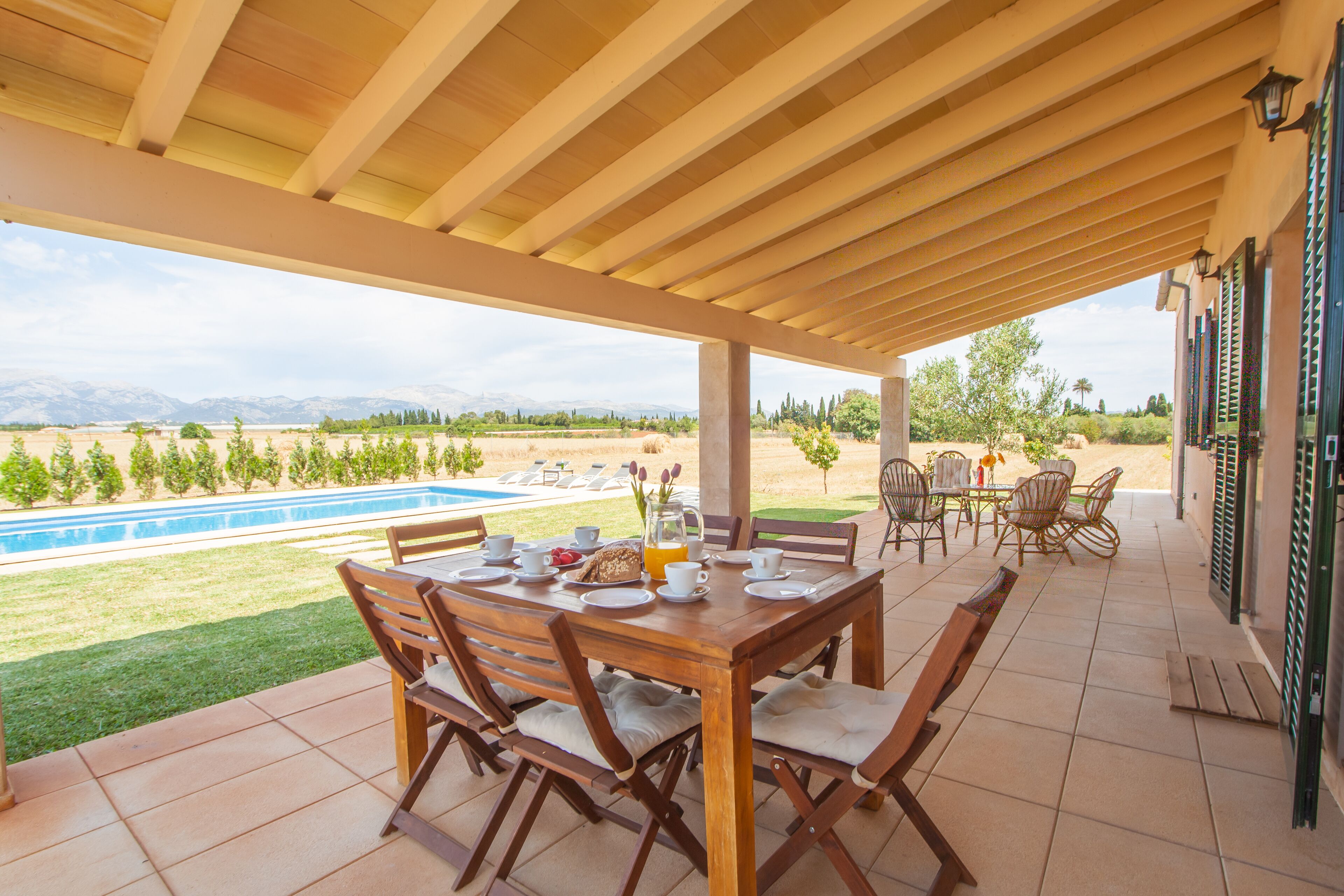 A covered patio with a wooden table set for breakfast, overlooking a swimming pool and a landscaped garden.