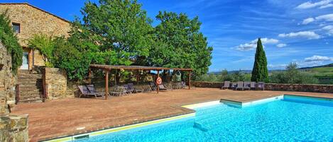 A swimming pool with a pergola and lounge chairs, surrounded by stone walls and trees.