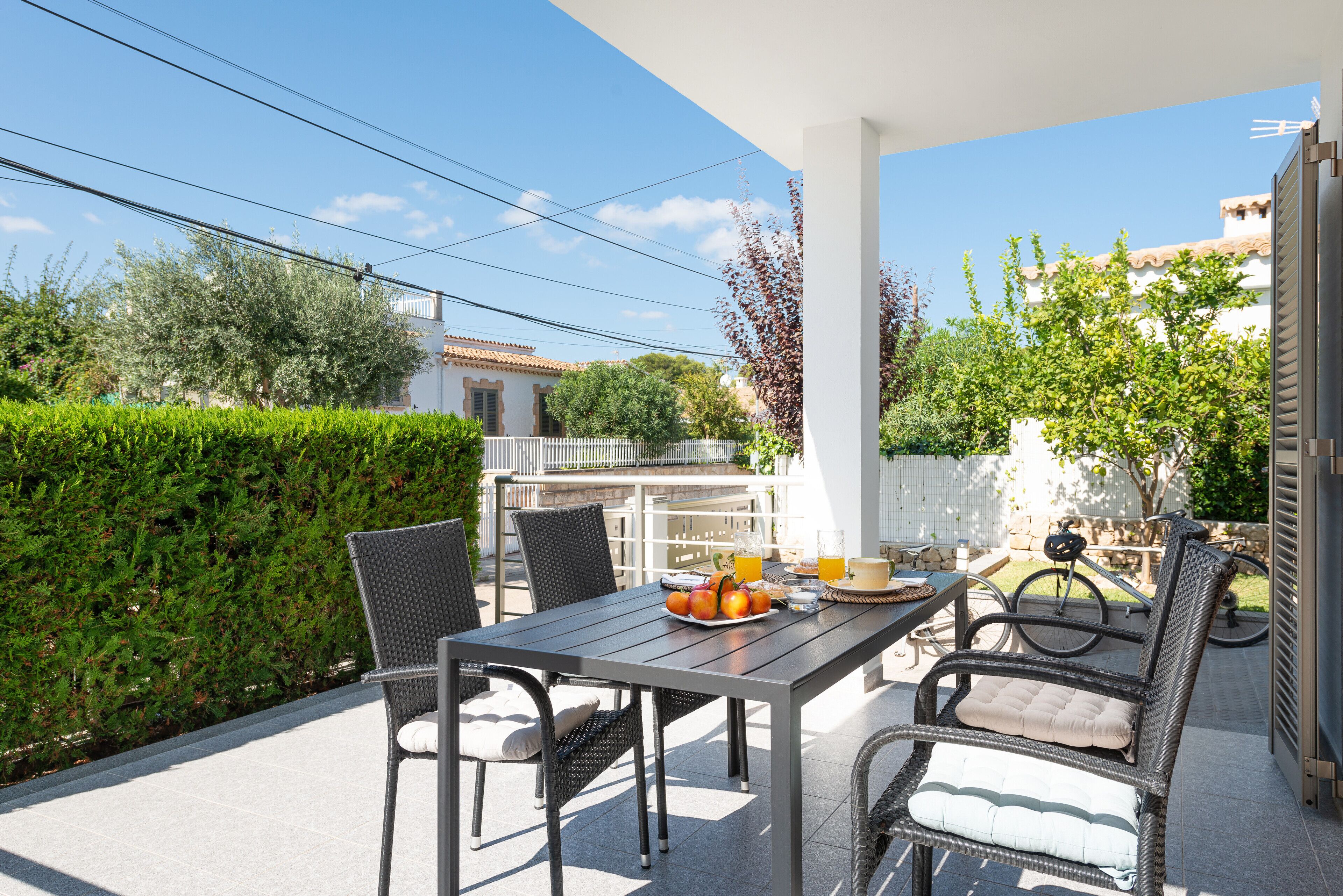 An outdoor patio with a table set for two, surrounded by wicker chairs, a bicycle, and a white fence.