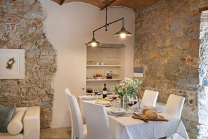 Un'area pranzo con parete in pietra, un tavolo bianco apparecchiato per quattro persone e una mensola con bicchieri e stoviglie sullo sfondo.