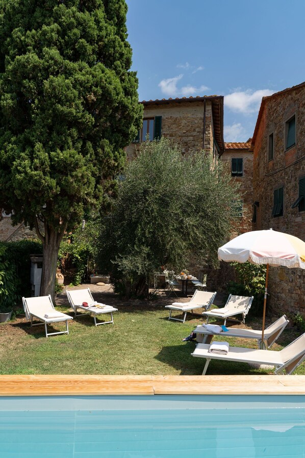 A poolside area with lounge chairs and an umbrella, surrounded by stone buildings and trees.