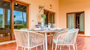 Un coin repas avec des chaises en osier et une table ronde dressée pour quatre personnes, une plante en pot et une bouteille de vin.