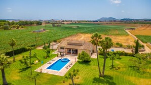 Une maison avec une piscine, entourée d’une végétation luxuriante et de champs.