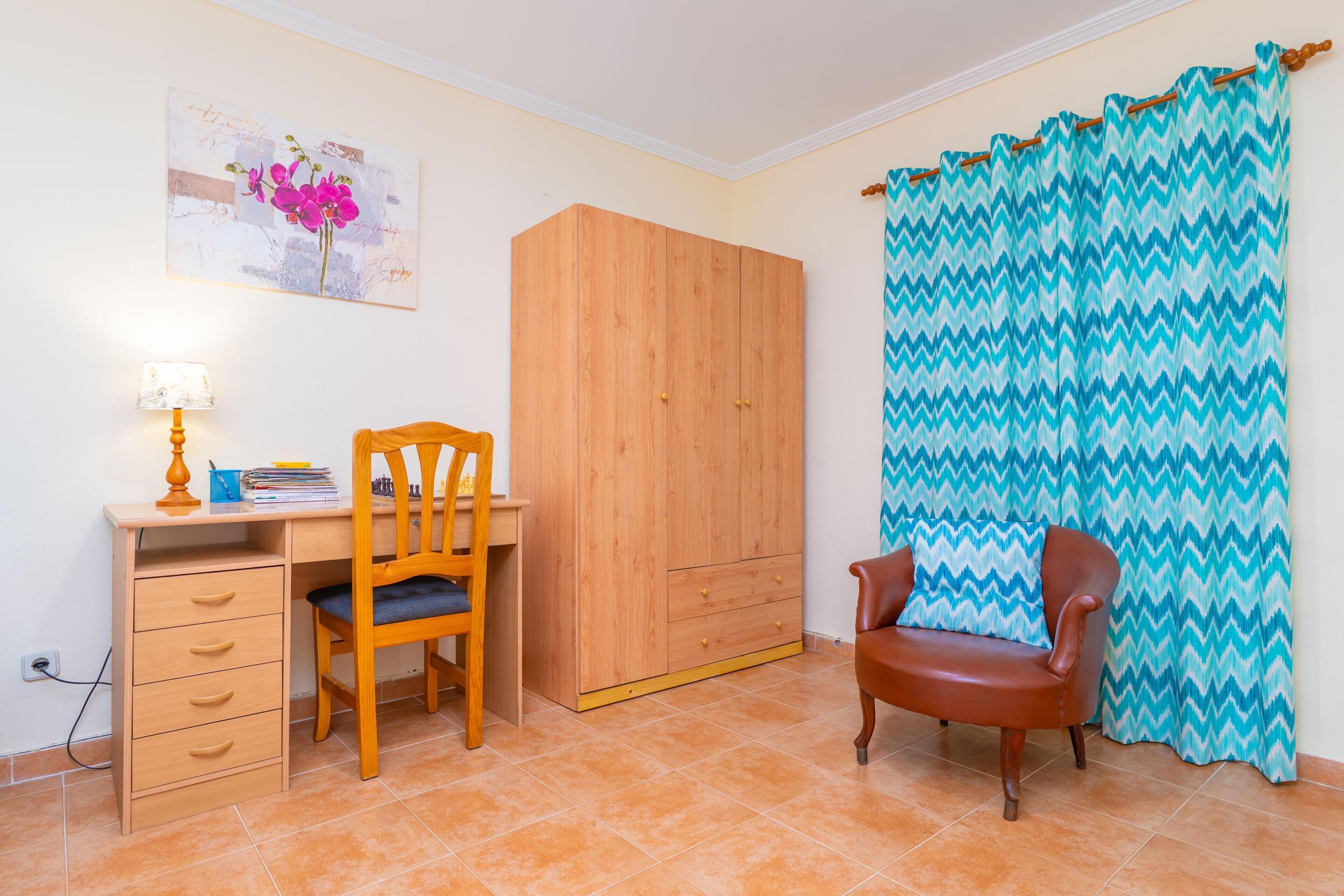 Een slaapkamer met een houten garderobe, een bureau met stoel, een bed met een blauw-wit ruit gordijn, en een schilderij aan de muur.