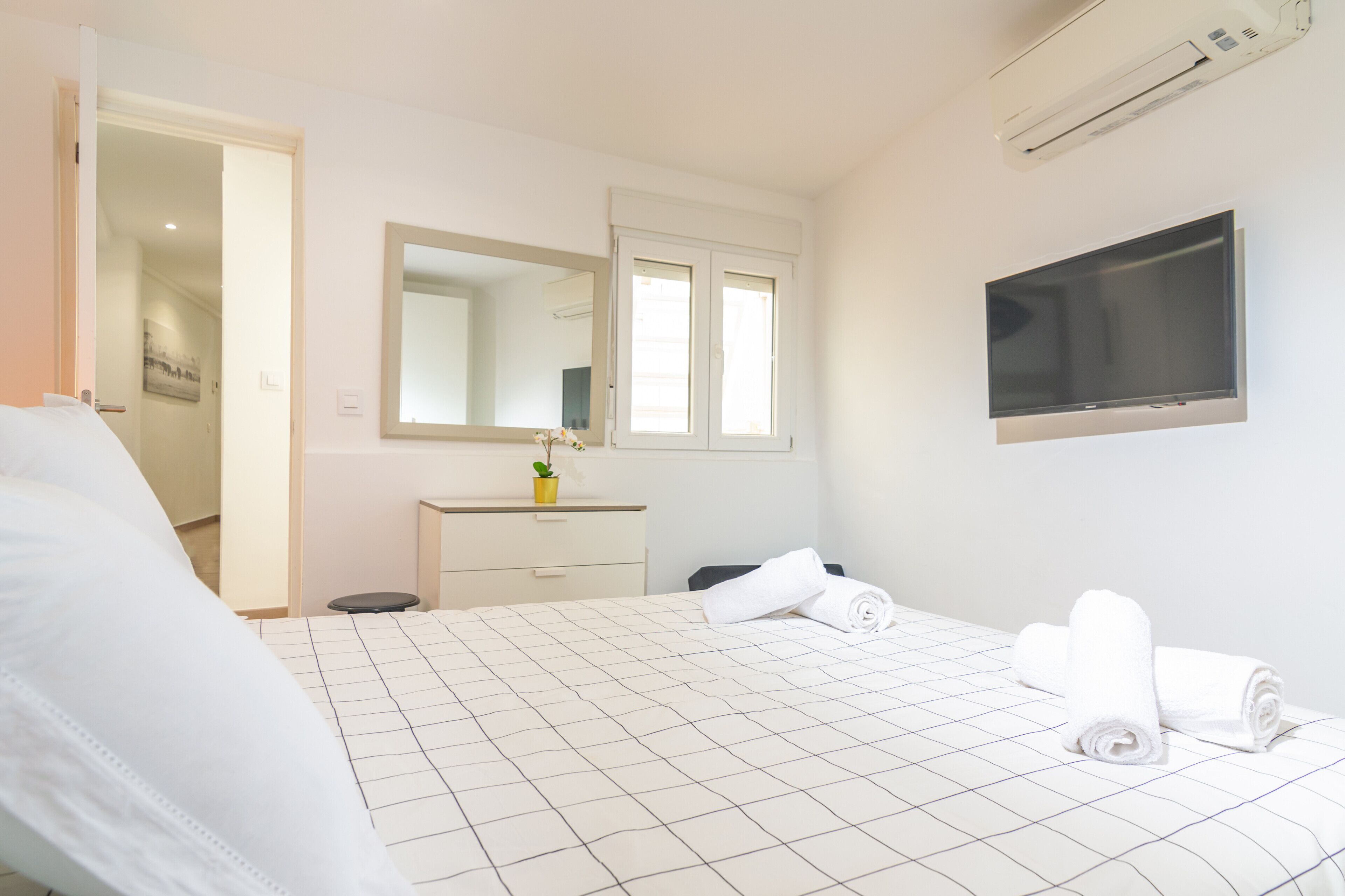 A hotel room with a bed, a TV mounted on the wall, a mirror above the dresser, and a door leading to another room.