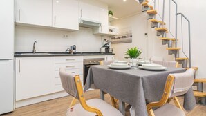 Eine moderne Küche mit weißen Schränken, einer eingebauten Mikrowelle und einer Spüle aus Edelstahl. Ein Essbereich mit Tisch für vier Personen, umgeben von Holzstühlen. Eine Treppe mit Metallgeländer führt zu einer oberen Ebene.