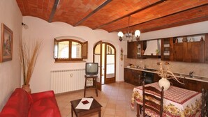 A cozy living room with a red sofa, a small table, and a TV. There's a kitchen with wooden cabinets and a dining area with a table and chairs. - Cosy apartment for 4 guests with WIFI, pool, TV and panoramic view, close to Montepulciano (Monticchiello)
