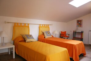 A room with two beds, each with an orange bedspread and matching pillows, a chair, a small table with a lamp, and a radiator.