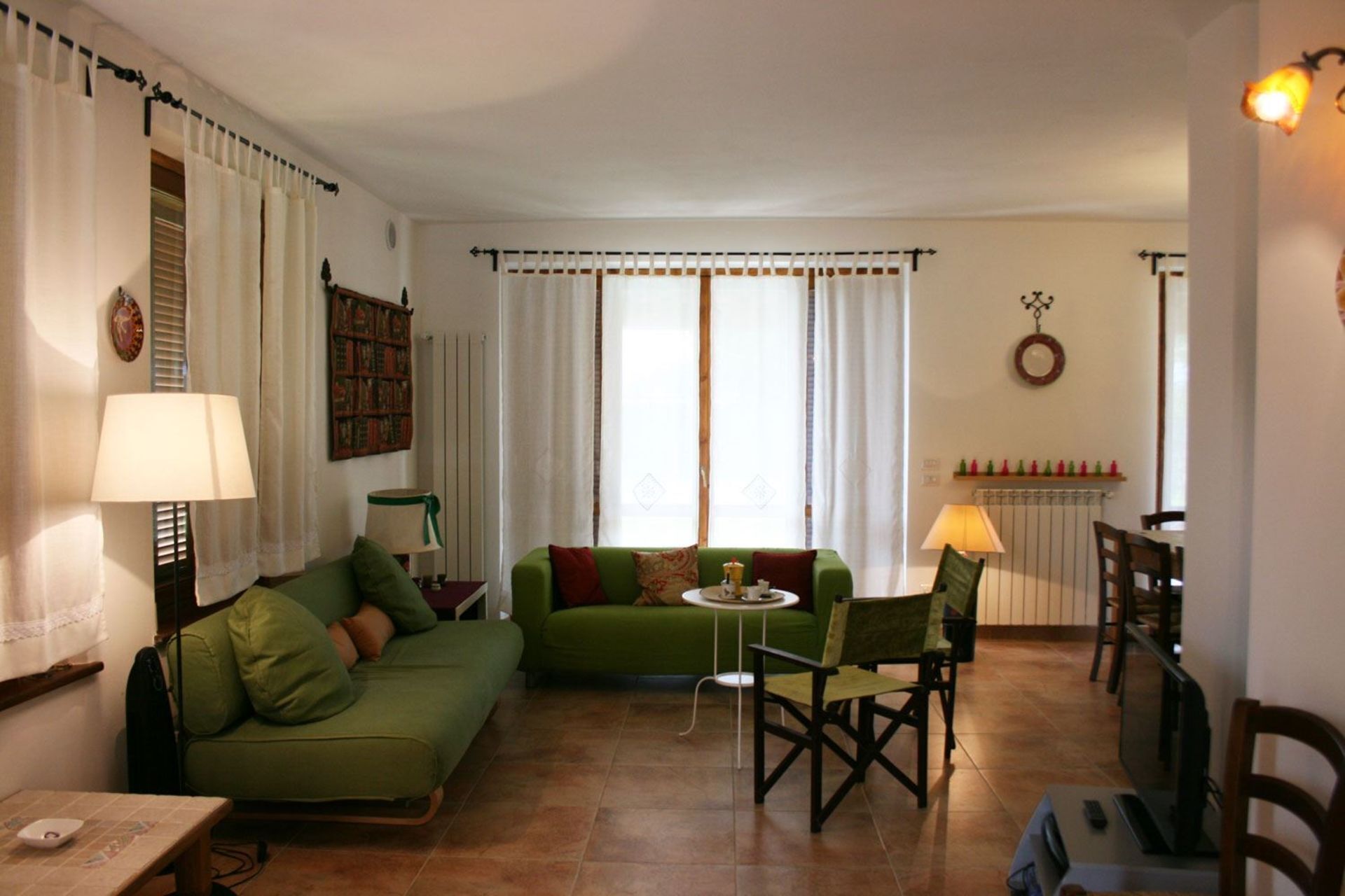 A living room with a green sofa, a small round table, and two chairs. There is a lamp on the left and a radiator on the right.