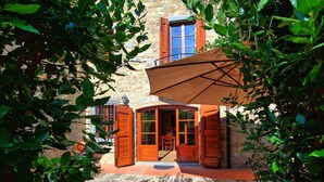 Un bâtiment en pierre avec une porte et des fenêtres en bois, un patio couvert avec une table et des chaises, et un chemin de pierre menant à l’entrée.