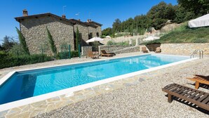 Une piscine aux bords recouverts de pierres, des chaises longues et un bâtiment en pierre doté d’une cheminée.