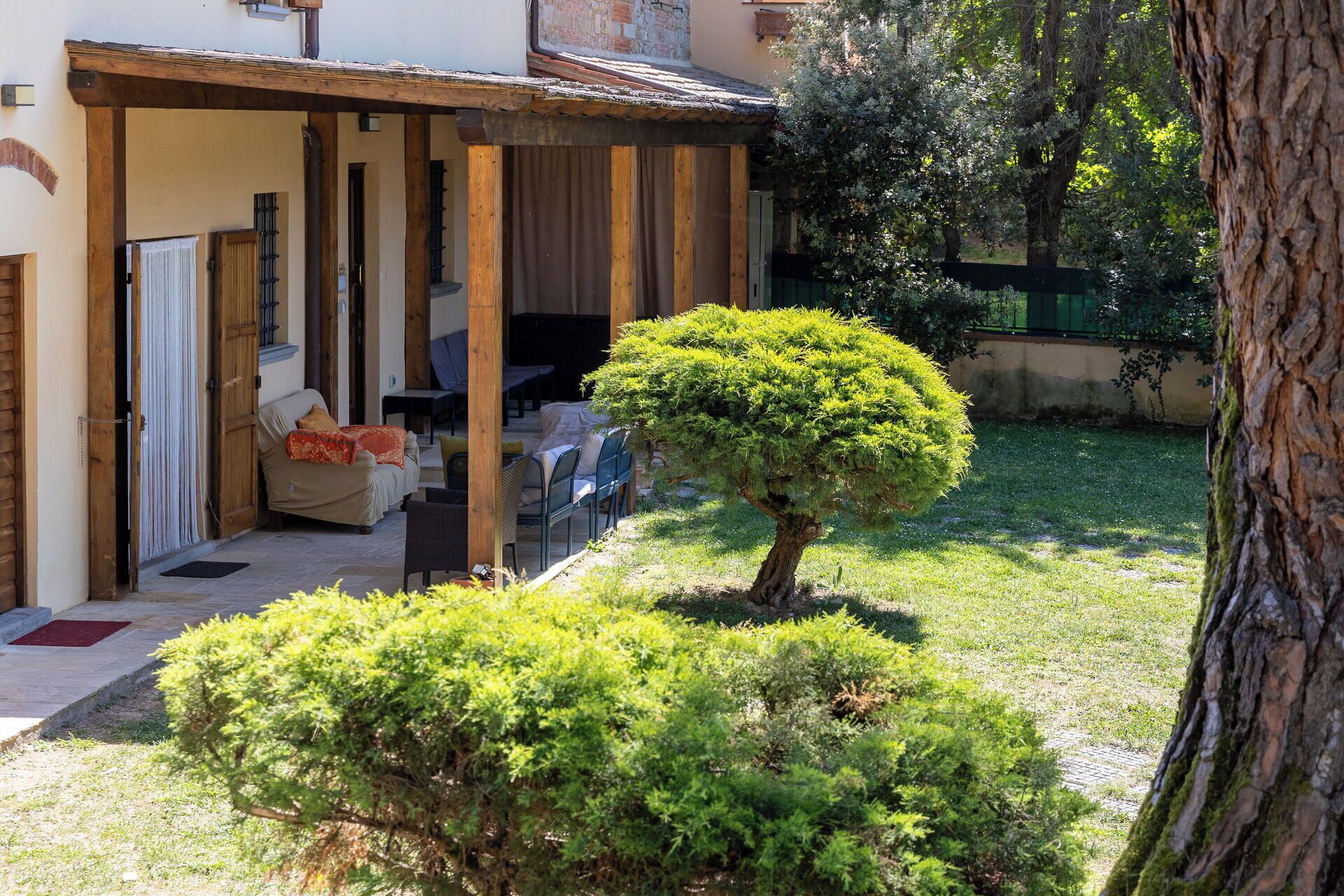 A covered outdoor seating area with a wooden pergola, a small tree, and a grassy lawn.