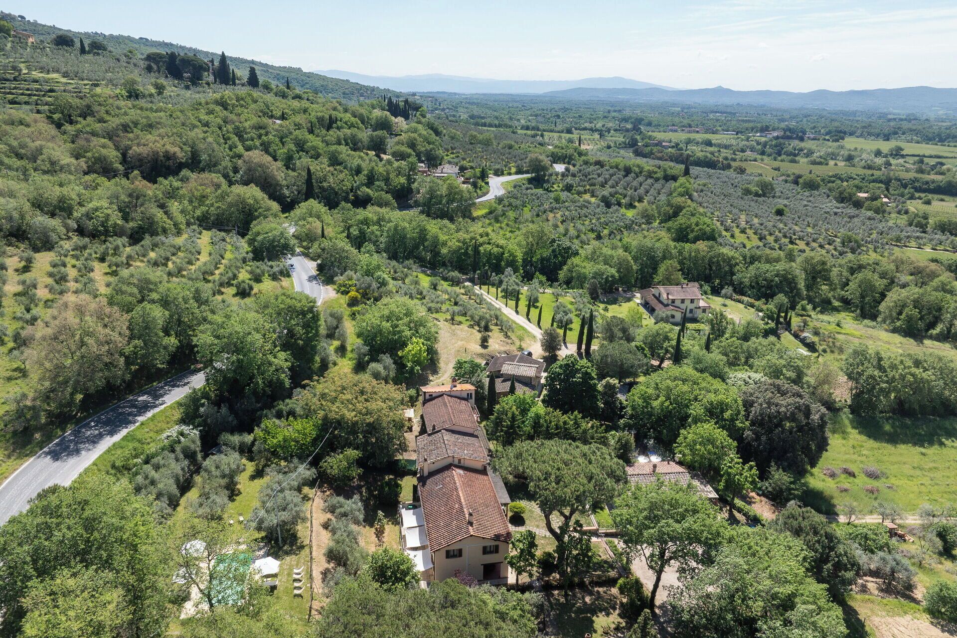 Paisatge rural amb un camí sinuós, una casa gran amb teulada de teules i vegetació al voltant.