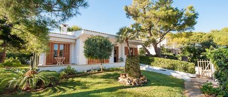 A house with a white exterior, wooden shutters, and a well-maintained garden with various trees and shrubs.
