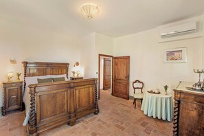 A bedroom with a wooden bed, a nightstand, a table with a lamp, a chair, and a door leading to another room.