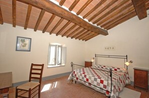 A bedroom with a wooden ceiling, a bed with a quilt, a chair, a bedside table, and a window.