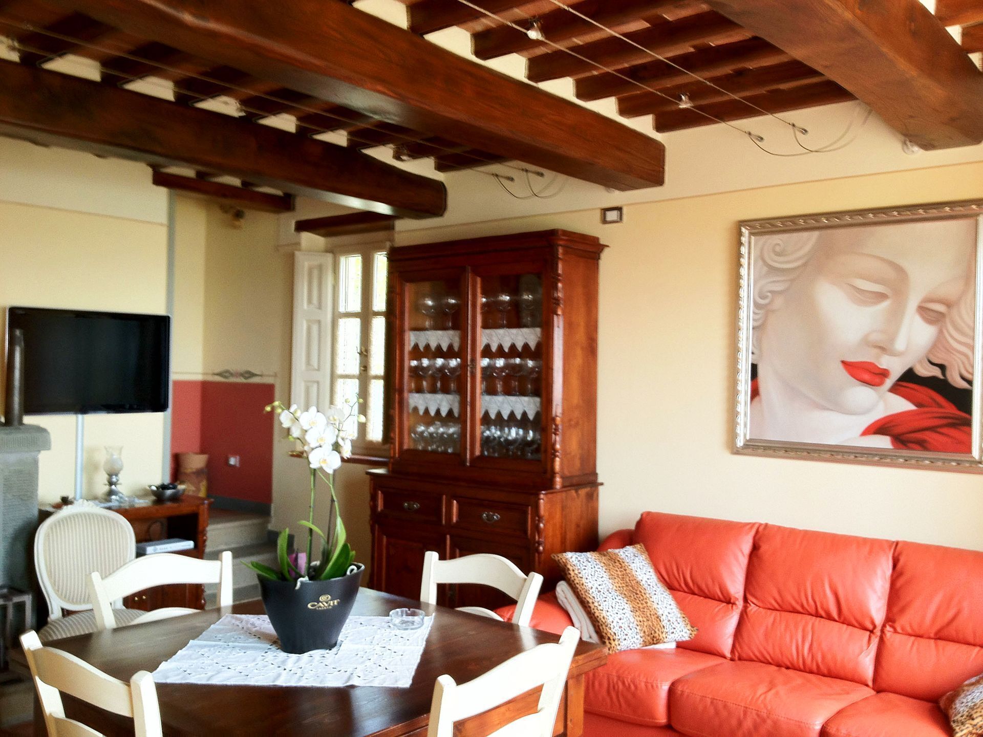 A living room with a wooden ceiling, a red sofa, a dining table with chairs, a cabinet with glassware, and a television on a stand.