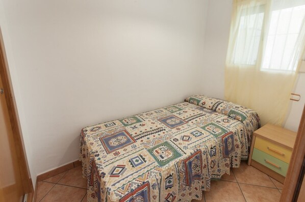 A small, single-bed room with a patterned bedspread, a wooden nightstand with drawers, and a window with sheer curtains.