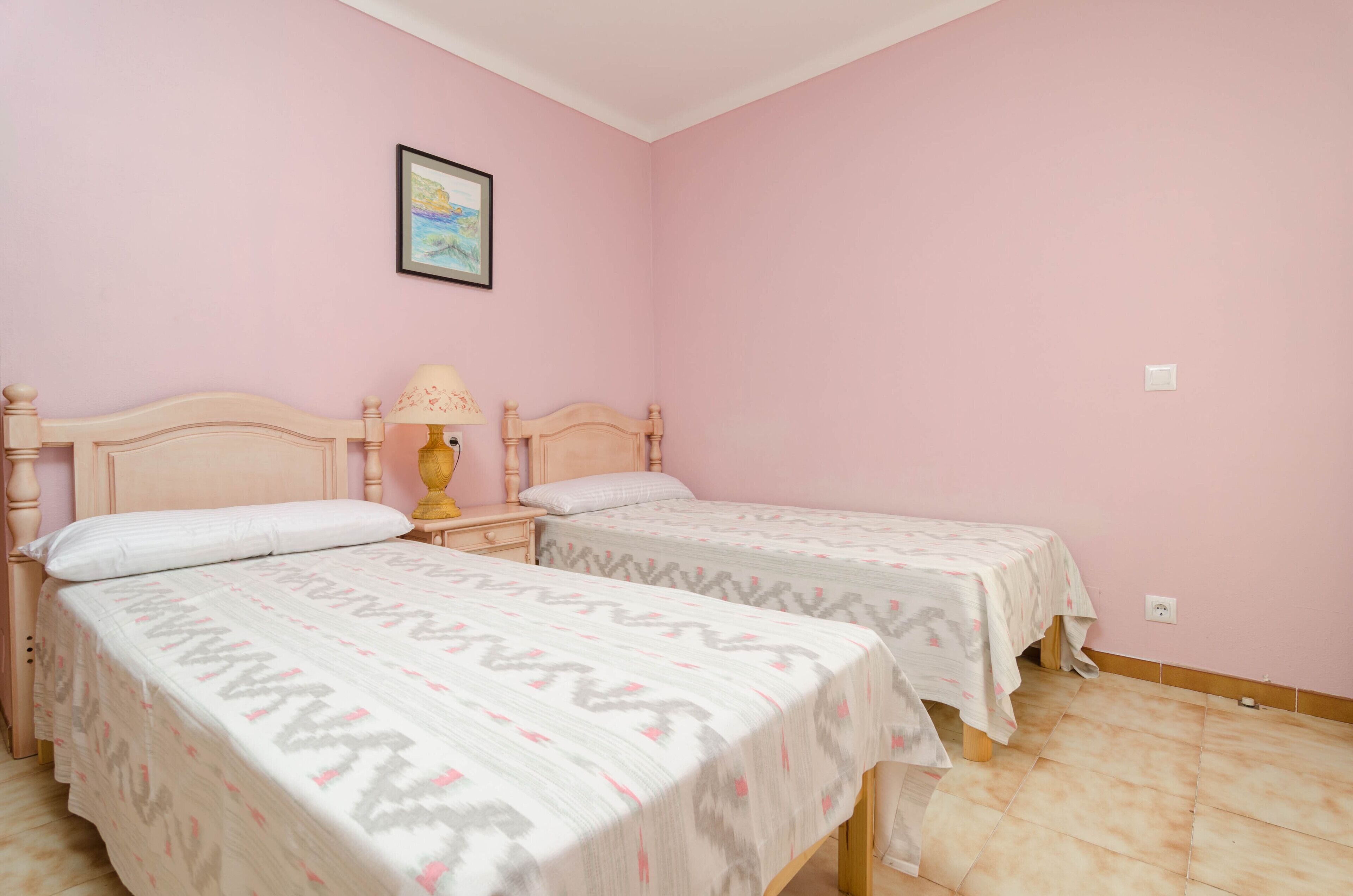 Een kamer met twee bedden, een nachtkastje met een lamp en een lijst met een afbeelding aan de muur.