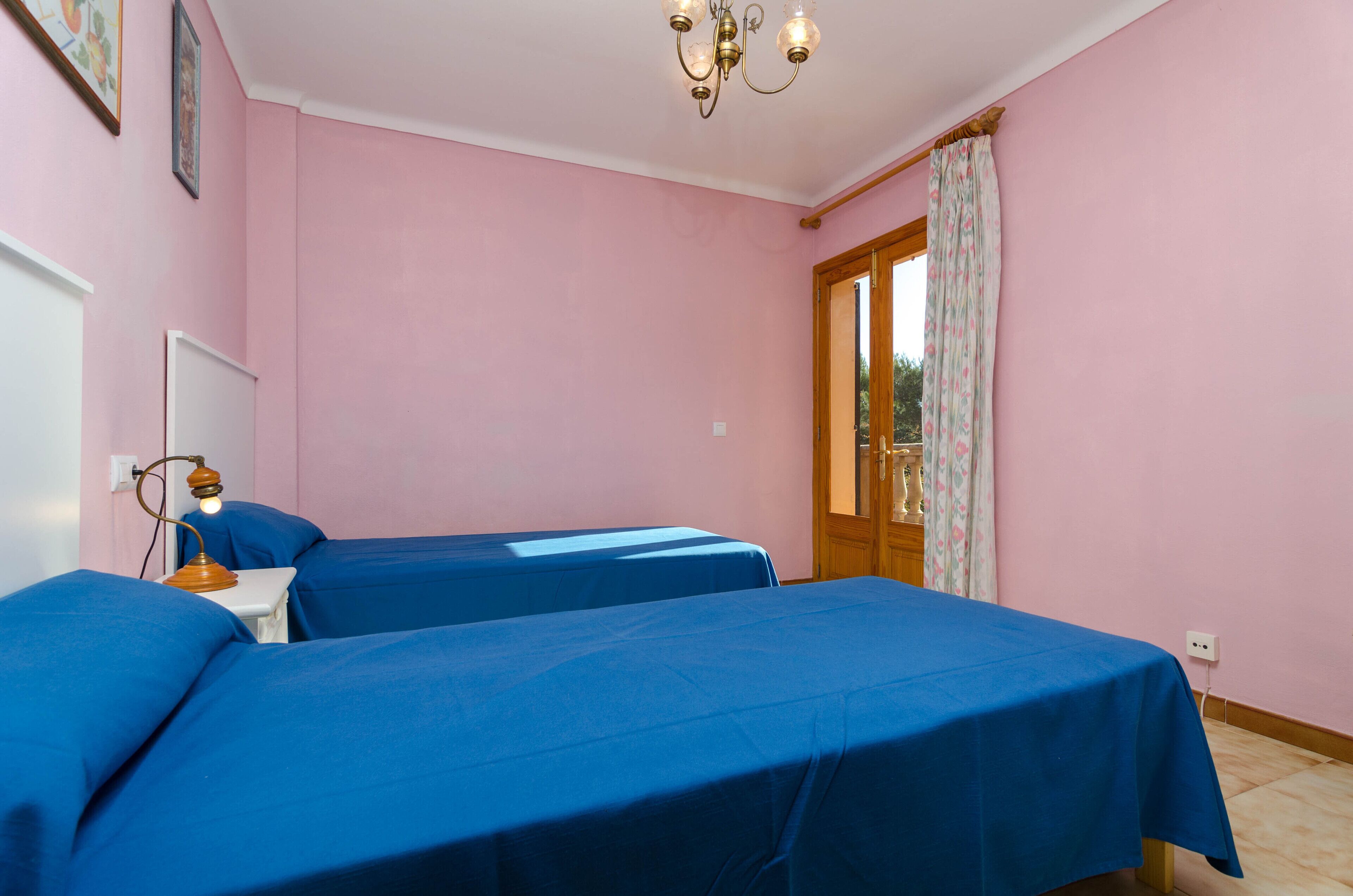 A bedroom with two beds, a wooden door, a window with floral curtains, and a chandelier.