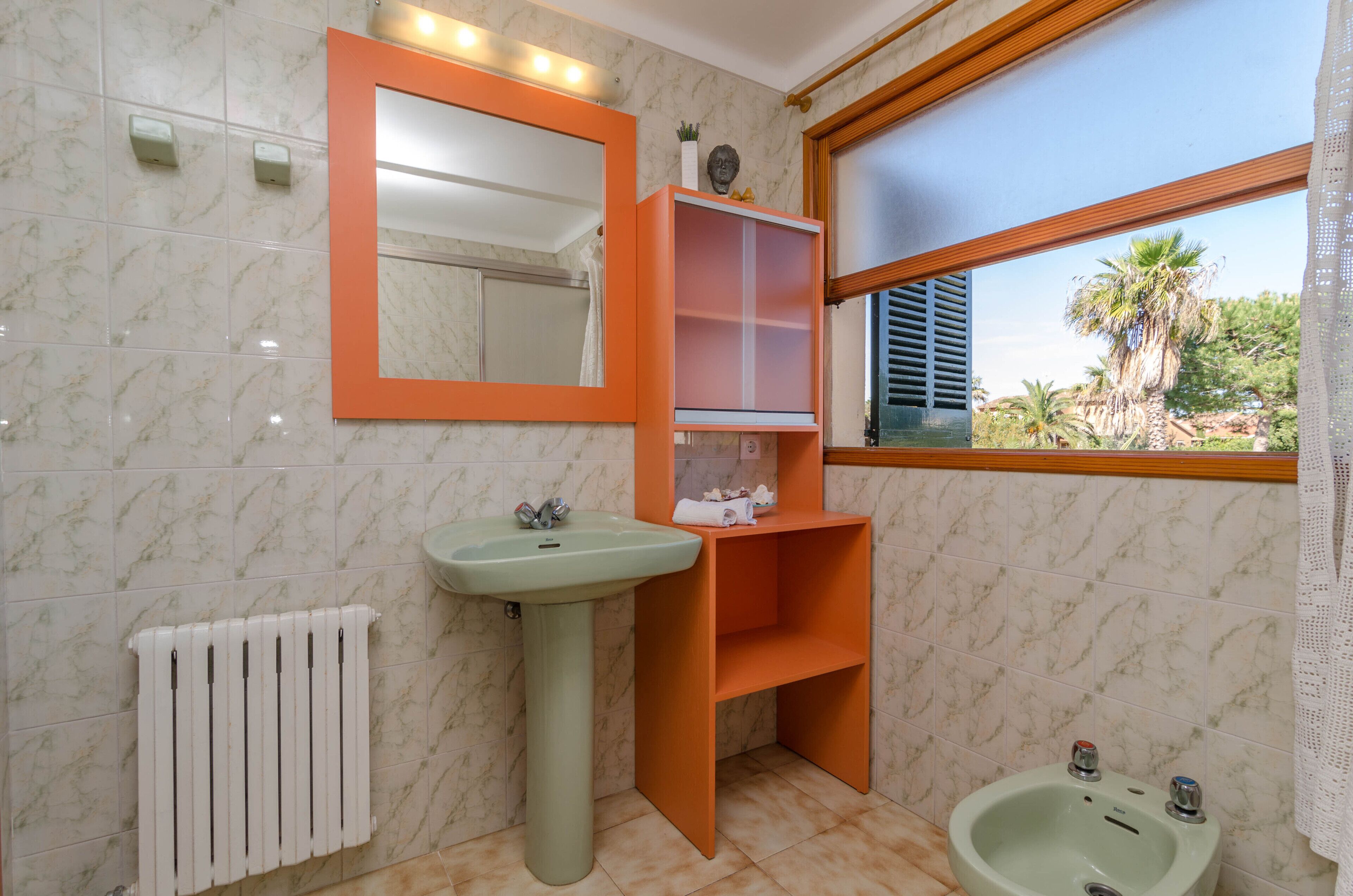 A bathroom with a pedestal sink, a mirror, a shelf unit, a radiator, and a window with a view of palm trees.