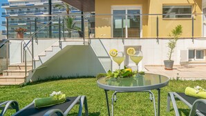 Uma área à beira da piscina com mesa de vidro, cadeiras de lounge e escadas que levam a uma varanda.