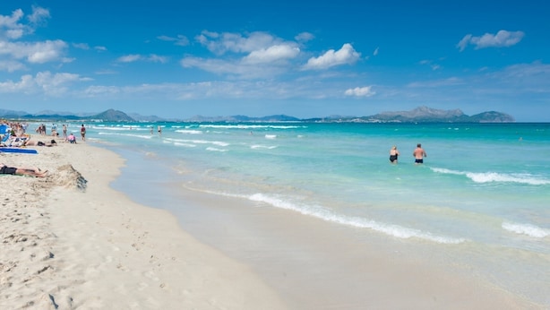 Uma praia com águas turquesa cristalinas, pessoas nadando e caminhando pela orla, e montanhas ao fundo.