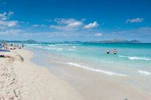 A beach with clear turquoise water, people swimming and walking along the shore, and mountains in the background.