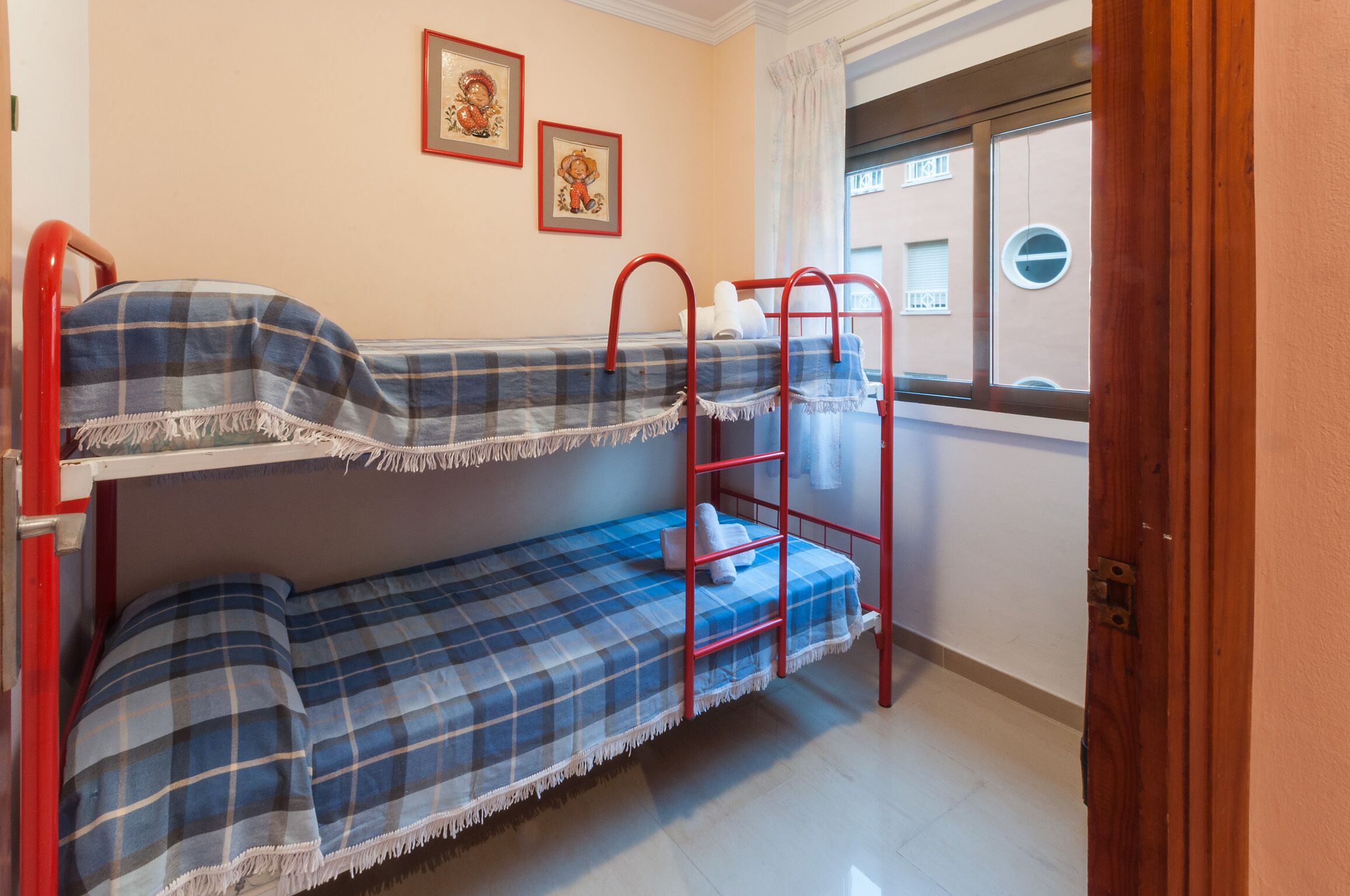 Une chambre avec un lit superposé, une couverture à carreaux, une fenêtre donnant sur des immeubles et deux tableaux encadrés accrochés au mur.