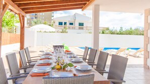 Un espace extérieur abrité, aménagé pour les repas, avec une longue table dressée, entouré de chaises en osier et donnant sur une piscine.