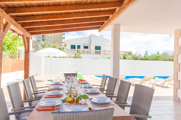 Uma área de refeições coberta ao ar livre com uma mesa comprida posta para uma refeição, rodeada por cadeiras de vime, com vista para uma piscina.