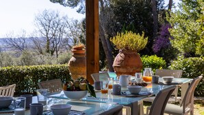 Un área de comedor al aire libre con una mesa preparada para el desayuno, rodeada de sillas y plantas en macetas.