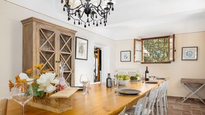 Un comedor con una mesa de madera dispuesta para seis comensales, sillas blancas, una araña y una ventana con vistas.