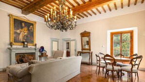 Una habitación con un sofá grande, sillas de madera, un candelabro, una chimenea y un ventanal amplio.