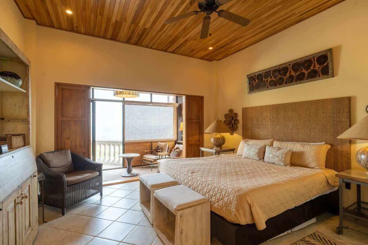 A spacious bedroom with a large bed, a ceiling fan, a wooden ceiling, a window with blinds, and a balcony with a chair and table.