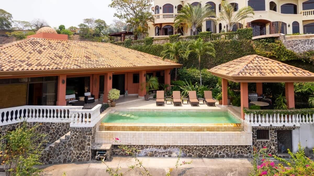 Um resort com piscina, espreguiçadeiras e uma escadaria de pedra que leva a um edifício com telhado ladrilhado.