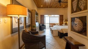 A hotel room with a wooden ceiling, a large window, a bed, a desk, and wicker furniture.