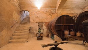 Une cave aux murs de pierre, avec un escalier en bois, une pompe à eau et des barils en bois.
