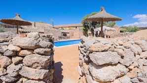 Un espace piscine entouré de murs en pierre, agrémenté de parasols, avec un ciel bleu dégagé et un bâtiment en arrière-plan.