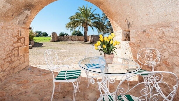 Un espace extérieur aménagé avec une arche en pierre, une table entourée de chaises et un pot de fleurs contenant des fleurs jaunes.