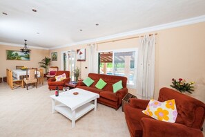 A spacious living room with a sofa set, a dining area, and a view through large windows.