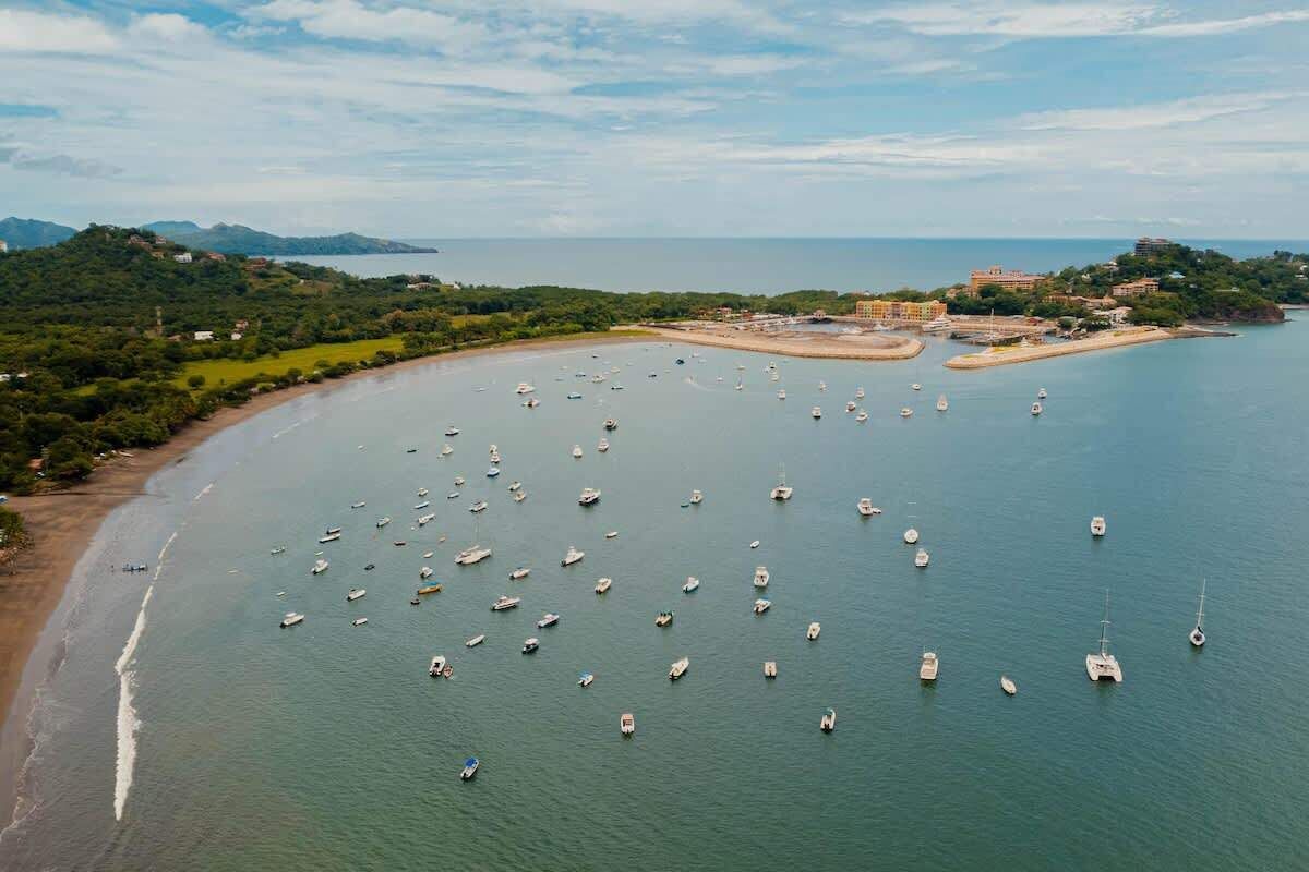 An aerial view of a coastal area with numerous boats anchored in the water, a sandy beach, and buildings on a hill in the background.