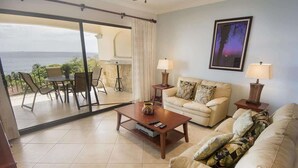 A living room with a sofa, coffee table, and a view of the ocean through a sliding glass door. - Oceanica 824- 2 Bedroom Ocean View Condo (Flamingo)